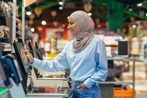 une femme acheteur dans une supermarché dans une hijab paie pour des biens à une en libre service vérifier, pratique un service pour les clients photo