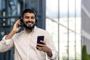 une Jeune Indien homme est permanent sur le rue dans une chemise et écouteurs, en portant une téléphone dans le sien mains, à la recherche à le filtrer, en parlant, écoute à musique, dansant photo