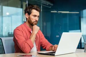 sérieux en pensant homme d'affaire proche en haut, homme dans rouge décontractée chemise travail à l'intérieur moderne bureau, mature patron travail avec portable à lieu de travail tandis que séance photo