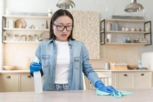 quarantaine moyens. une Jeune magnifique asiatique femme désinfecte le loger, lingettes le table avec une tissu dans bleu caoutchouc gants, Vaporisateurs le produit. virages de saleté et poussière. photo