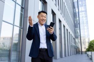 content Jeune asiatique homme homme d'affaire dans costume permanent près Bureau bâtiment avec téléphone et sous le choc content succès, diffusion bras. photo