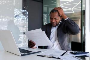 frustré et en colère financier à lieu de travail à l'intérieur bureau, en colère patron révision factures contrats et rapports séance à table avec ordinateur portable, homme d'affaire derrière papier travail. photo