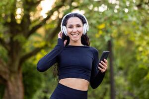 portrait de de bonne humeur femelle athlète dans serré raccord tenue de sport écoute à la musique dans écouteurs connecté à mobile téléphone. frisé Dame pratiquant en bonne santé mode de vie et profiter exercice sur ouvert air. photo