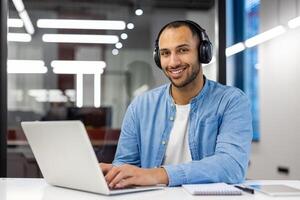 portrait de une souriant Jeune homme séance dans le Bureau avec une portable dans écouteurs et à la recherche en toute confiance dans le caméra. photo