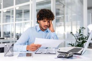 dérangé financier à la recherche à Ventes et revenu graphique de entreprise, homme d'affaire à l'intérieur Bureau déçu avec Ventes résultats et réalisations, hispanique homme séance à table avec les documents et portable. photo