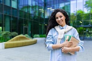 portrait de une Jeune magnifique hispanique femelle étudiant, femme à l'extérieur une Université Campus souriant et à la recherche à le caméra, en portant cahiers d'exercices, livres et manuels pour en étudiant. photo