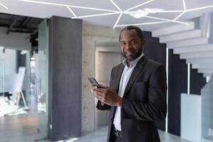 portrait de un plus âgée Afro-américain homme dans une costume permanent dans le couloir de un Bureau centre, bavardage sur le téléphone, attendre pour un rendez-vous, souriant à le caméra. photo