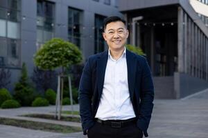 portrait de une souriant asiatique homme d'affaire dans une costume permanent en toute confiance à l'extérieur une contemporain Bureau complexe sur une ensoleillé journée. photo