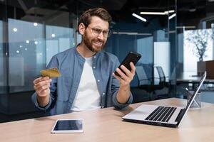 frustré et trompé homme d'affaire dans Bureau en essayant à faire achat dans en ligne boutique et en ligne argent transfert de banque crédit carte, agacé homme travail à l'intérieur Bureau en utilisant portable et téléphone. photo