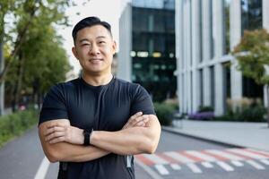 portrait de une souriant Jeune asiatique Masculin athlète permanent sur une ville rue, à la recherche à le caméra avec le sien bras franchi. photo