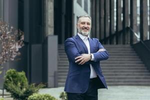 portrait de une réussi Sénior Masculin homme d'affaire dans une affaires costume permanent en toute confiance près le Bureau centre dans plein hauteur, bras franchi, souriant. photo