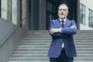 portrait de une Sénior aux cheveux gris homme directeur, PDG sur le Contexte de le escaliers de le Bureau centre. il est permanent sérieux dans une costume, bras franchi dans devant, à la recherche à le côté. photo