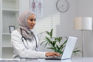 une musulman femelle médecin dans hijab, souriant tandis que travail sur sa portable dans une moderne clinique bureau. le scène reflète une professionnel et réconfortant soins de santé environnement. photo