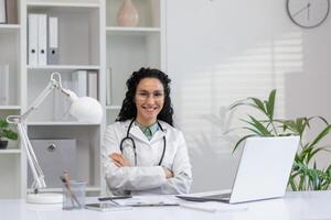 professionnel hispanique femelle médecin avec bras franchi séance à sa bureau avec une portable et stéthoscope dans bureau. photo