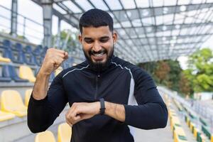 une barbu Jeune homme dans tenue de sport fête le sien aptitude objectif avec une poing pompe à un Extérieur stade. photo