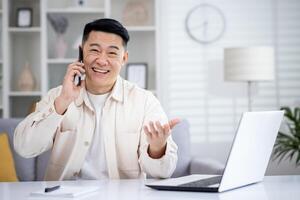 asiatique Jeune souriant Masculin pigiste travail à maison, séance à le table et parlant émotionnellement sur le mobile téléphone tandis que faire des gestes à le caméra. photo