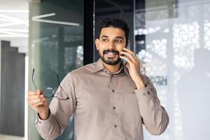 de bonne humeur Jeune homme d'affaire parlant sur une téléphone intelligent avec des lunettes dans main, permanent dans une contemporain Bureau paramètre. photo