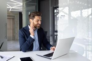 inquiet Jeune homme dans affaires costume travail dans Bureau à ordinateur portable, séance renfrogné et en portant main à joue. il se sent une fort mal aux dents. photo