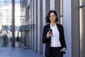 une Jeune affaires femme est en marchant vers le bas le rue près le Bureau centres dans une affaires costume et en utilisant une mobile téléphone. photo