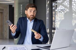 portrait de une Jeune homme d'affaire séance dans le Bureau à le bureau, en portant une mobile téléphone et une crédit carte dans le sien mains, à la recherche déçu et confus à le caméra. photo