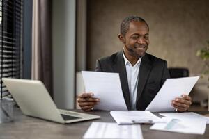 une professionnel dans une costume soigneusement analyses documents, entouré par une portable et rapports sur une bureau dans une bien éclairé bureau. photo