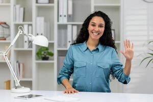 amical femme souriant et agitant à le caméra pendant une décontractée appel de une Accueil Bureau paramètre. photo