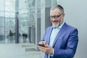 content Sénior Beau aux cheveux gris homme, homme d'affaire dans costume et lunettes. détient le téléphone dans le sien mains, reçu bien nouvelles, lit le message. il des stands sur le rue près une moderne bâtiment photo