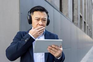 une Jeune asiatique homme dans une costume et écouteurs est permanent à l'extérieur et est à la recherche terrifié à le écran de le tablette il est en portant dans le sien mains, ayant reçu et révision mal nouvelles. photo