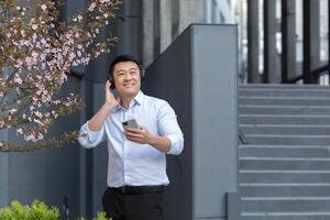 satisfait Jeune Beau asiatique homme dans blanc chemise détient téléphone et écoute à livre audio, Podcast dans noir écouteurs. en marchant sur le rue près une moderne bâtiment et une magnifique magnolia arbre. photo