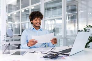 réussi financier comptable travail à l'intérieur Bureau avec portable et documents, homme satisfait avec Audit résultats souriant et en train de lire financier rapports, homme d'affaire en utilisant portable pour formalités administratives. photo