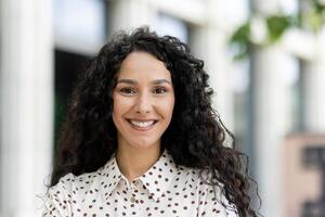 portrait de une radiant femme d'affaires avec frisé cheveux souriant en toute confiance, permanent en plein air avec un Urbain toile de fond. photo