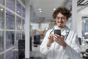 une homme dans une blanc laboratoire manteau et stéthoscope détient une cellule téléphone à l'intérieur une médical Bureau de une clinique. photo