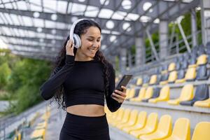 une souriant femme écoute à la musique par blanc écouteurs, en utilisant sa téléphone intelligent dans un vide stade sièges zone. photo