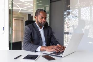 sérieux mature homme d'affaire travail sur portable à l'intérieur Bureau à lieu de travail, africain américain patron réfléchi dans affaires costume, réussi expérimenté investisseur banquier. photo