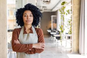 portrait de une sérieux et confiant Afro-américain femme d'affaires, propriétaire de une restaurant, bar, permanent avec sa bras franchi et à la recherche à le caméra. photo