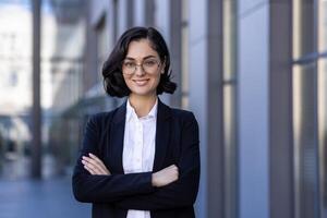 fermer portrait de une Jeune femme d'affaires permanent à l'extérieur un Bureau bâtiment avec sa bras franchi plus de sa poitrine. en toute confiance et en souriant à la recherche à le caméra. photo