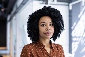 une Jeune africain américain femme d'affaires avec frisé cheveux, portant une polka point chemisier, des stands en toute confiance dans une bien éclairé moderne Bureau environnement, exsudant professionnalisme et équilibre. photo