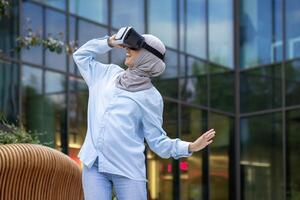 une joyeux musulman femme avec une hijab jouit en utilisant une virtuel réalité casque, exprimer excitation en plein air avec moderne architecture Contexte. photo