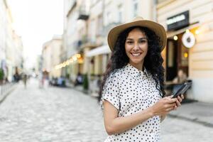 portrait de une Jeune magnifique Latin américain femme en marchant dans le soir ville, en portant une téléphone, souriant et à la recherche à le caméra. photo