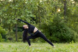 un actif Jeune femme effectue une yoga pose en plein air, entouré par vibrant vert des arbres et le lueur de le le coucher du soleil. photo