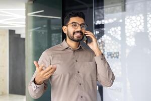 une homme est parlant sur le sien cellule téléphone dans une pièce avec une verre mur photo