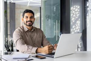 concentré Indien homme d'affaire en utilisant une portable à le sien bureau dans une brillant contemporain Bureau paramètre, avec formalités administratives et numérique tablette. photo