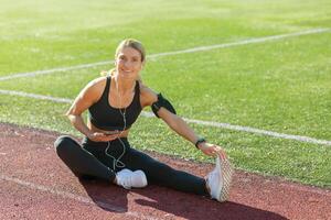 en forme femelle athlète séance sur une des sports Piste élongation sa jambe et en utilisant sa téléphone, écouteurs dans, pendant faire des exercices Pause. photo