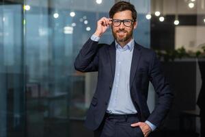 portrait de réussi mature chef, Sénior homme d'affaire dans des lunettes et affaires costume à la recherche à caméra et souriant, homme travail à l'intérieur moderne Bureau bâtiment. photo