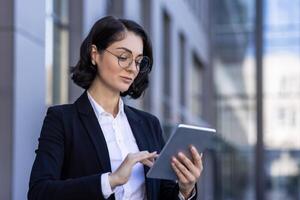 une Jeune femelle avocat est permanent à l'extérieur le tribunal dans une affaires costume et des lunettes et est sérieusement en utilisant une tablette. photo