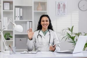 une amical femelle médecin portant une laboratoire manteau avec une stéthoscope sourit et vagues dans une brillant clinique bureau, exsudant professionnalisme et chaleur. photo