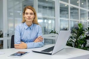 une concentré professionnel femme est assis à sa Bureau bureau, travail sérieusement sur une moderne portable entouré par Bureau essentiel. photo