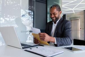 africain américain homme d'affaire en train de lire bien nouvelles, homme à l'intérieur Bureau lieu de travail en portant courrier enveloppe lettre avec bien nouvelles, patron dans affaires costume mature et adulte. photo