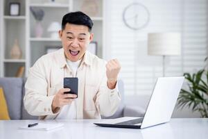 le gagnant est séance à le table à maison, le homme est content à recevoir le en ligne notification de le la victoire et Succès sur le téléphone, le asiatique homme est en portant le sien main en haut et en portant téléphone intelligent. photo