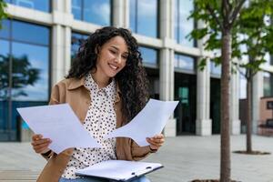 une jeune, de bonne humeur professionnel femme est permanent en plein air, soigneusement examiner papiers avec une sourire sur sa affronter. le Contexte Caractéristiques un Bureau bâtiment, signalisation une entreprise environnement. photo